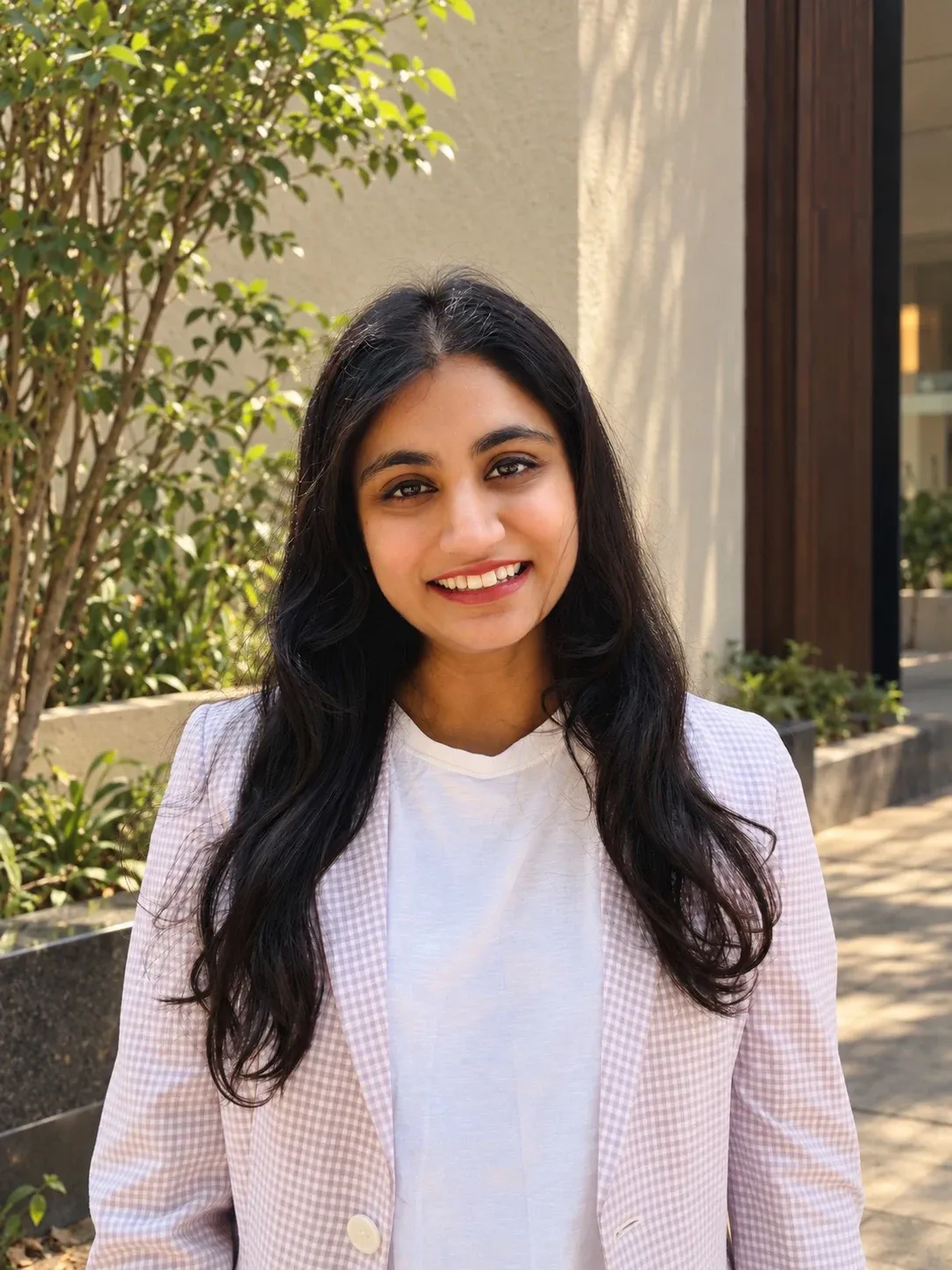 Nanditha smiling outdoors in a white top and a light checked blazer, beside a leafy plant.